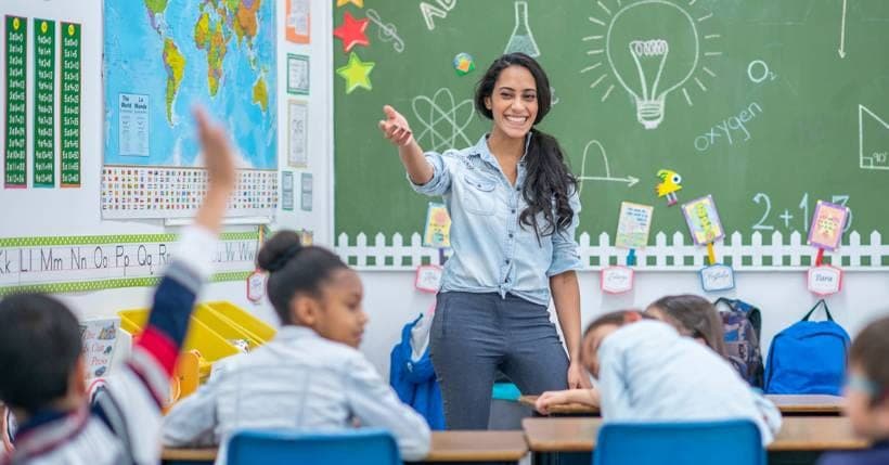 Teacher pointing to a lightbulb drawing on a classroom whiteboard while young students raise their hands and take notes